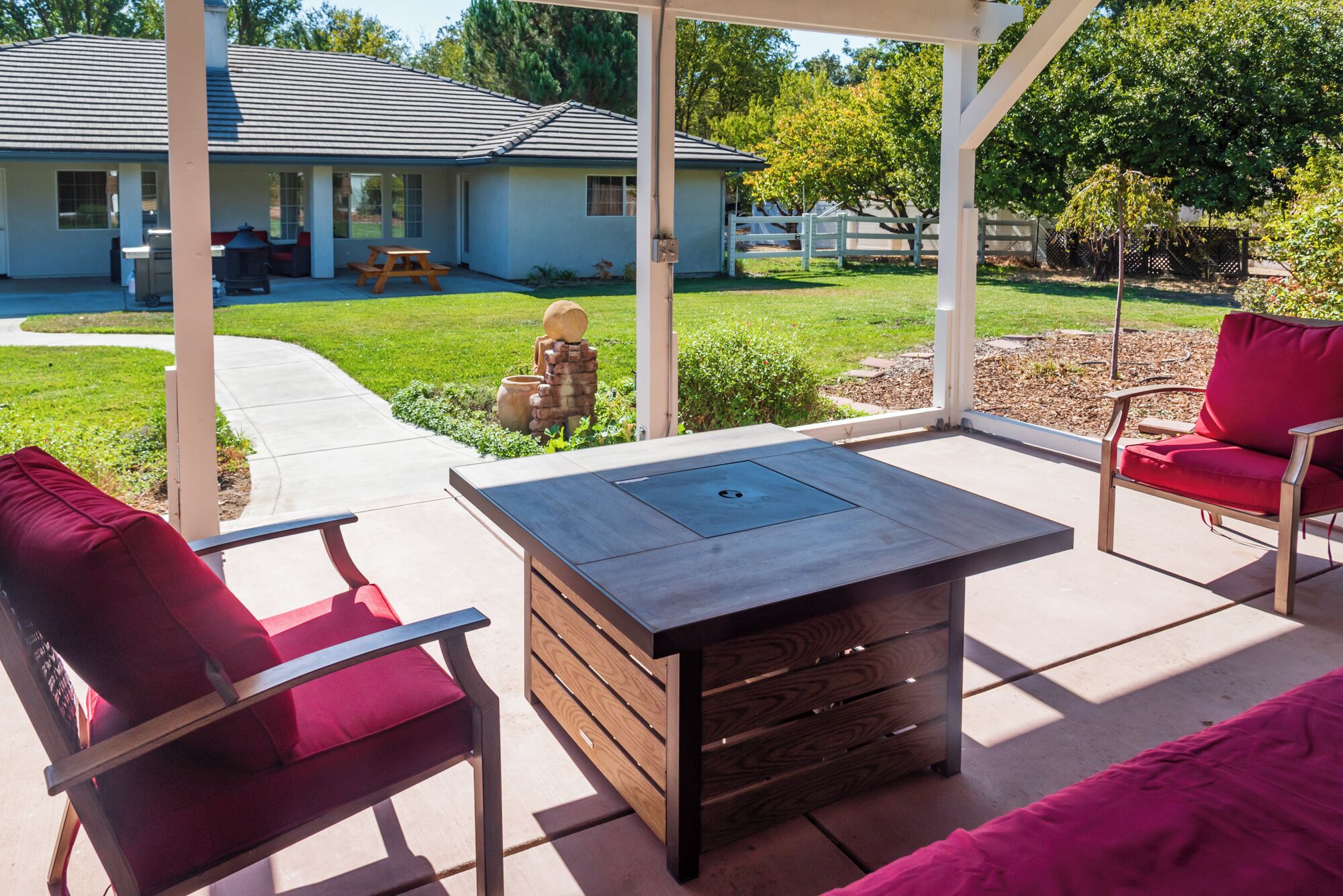 An outdoor patio with a built-in fire pit table, red cushioned chairs, and a view of a house in the background.