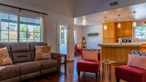 Een ruime woonkamer met een bruine bank en rode fauteuils, een houten vloer en een keuken met houten kastjes en hanglampen.