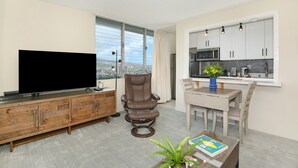 A modern living room with a flat-screen TV, a brown leather chair, a dining table with chairs, and a kitchen area with cabinets and a microwave. - HIGH Floor w/Canal, Mtn and Partial Ocean Views! AC, W/D, WiFi, Pool,Parking! (Honolulu)