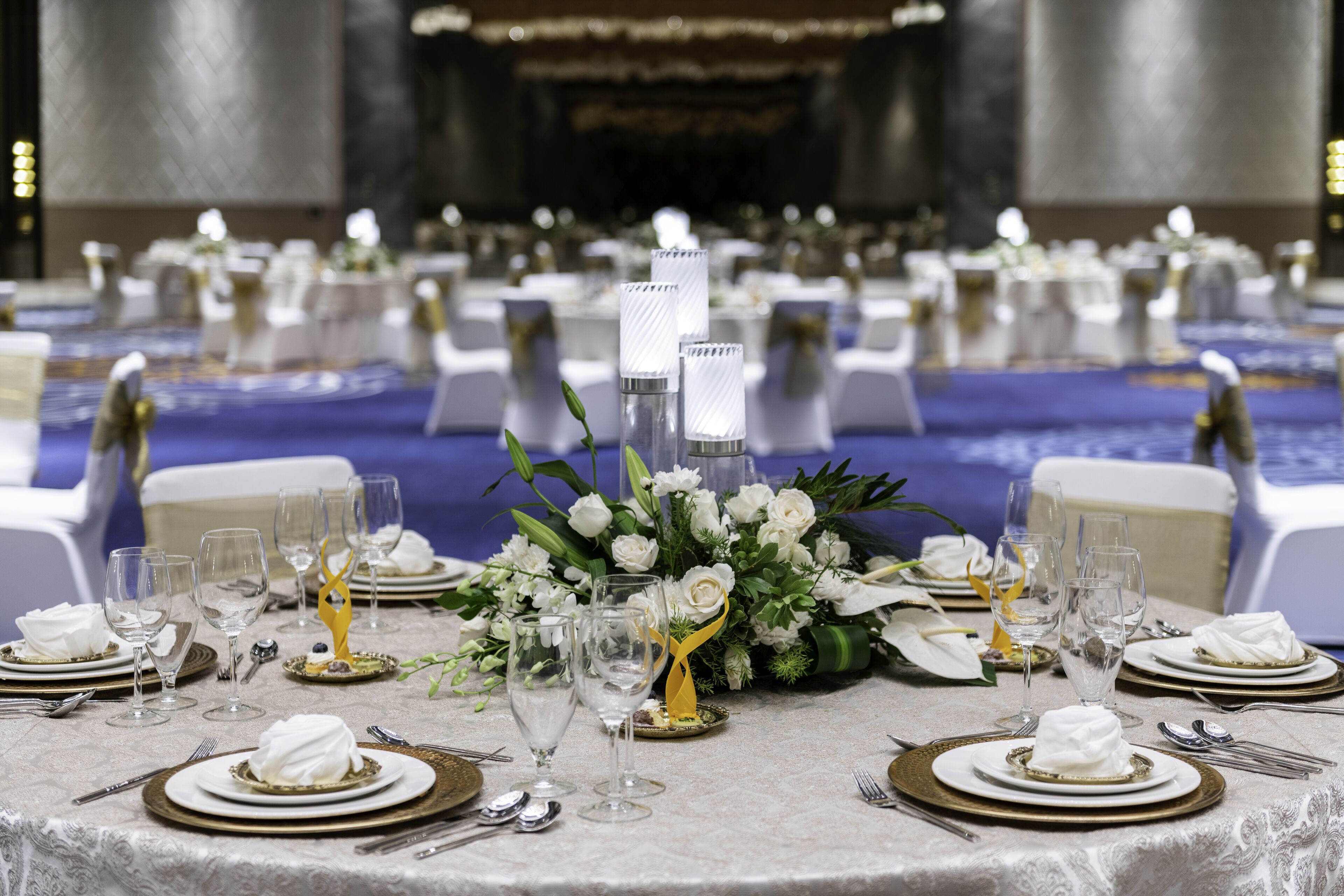 Un salón de banquetes con mesas redondas dispuestas para un evento formal, con manteles blancos, platos con borde dorado y cristalería transparente.