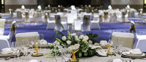 Un salón de banquetes con mesas redondas dispuestas para un evento formal, con manteles blancos, platos con borde dorado y cristalería transparente.