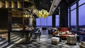 A modern bar area with a high bar counter, seating with a red sofa and ottoman, and a large window offering a city view. - Taj Lakefront Bhopal (Bhopal)