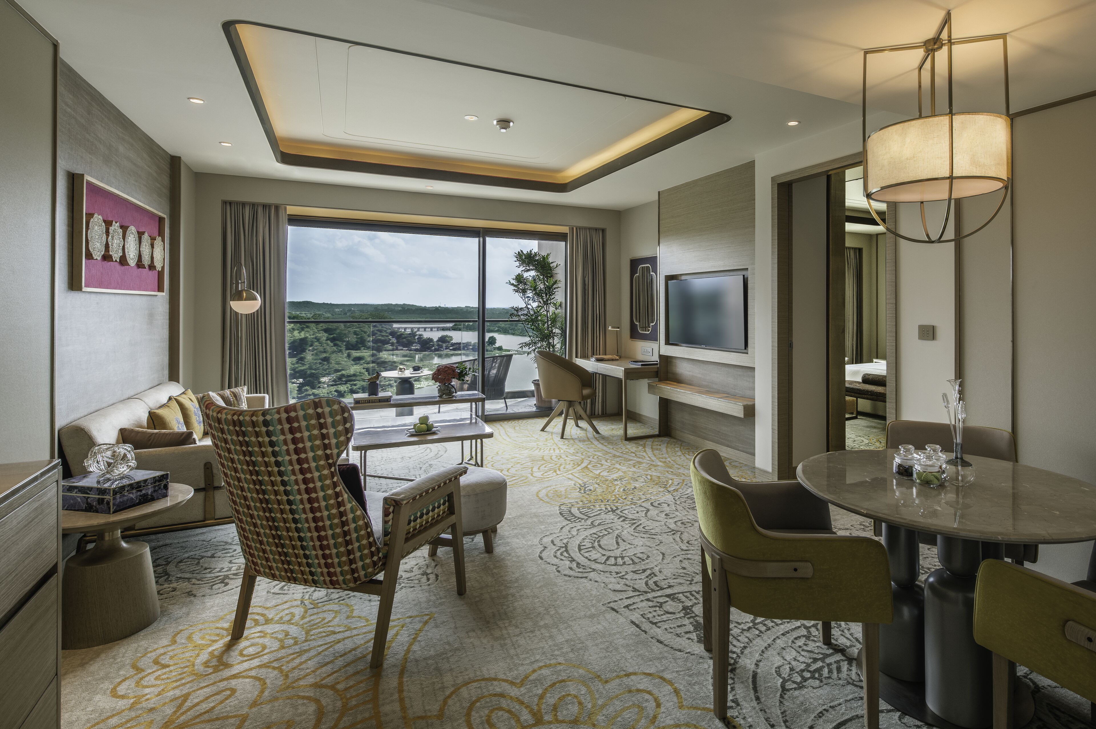 A modern hotel room with a large window offering a view of the outdoors, a dining area with a round table, and a seating area with a sofa and chairs.