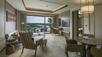 A modern hotel room with a large window offering a view of the outdoors, a dining area with a round table, and a seating area with a sofa and chairs.