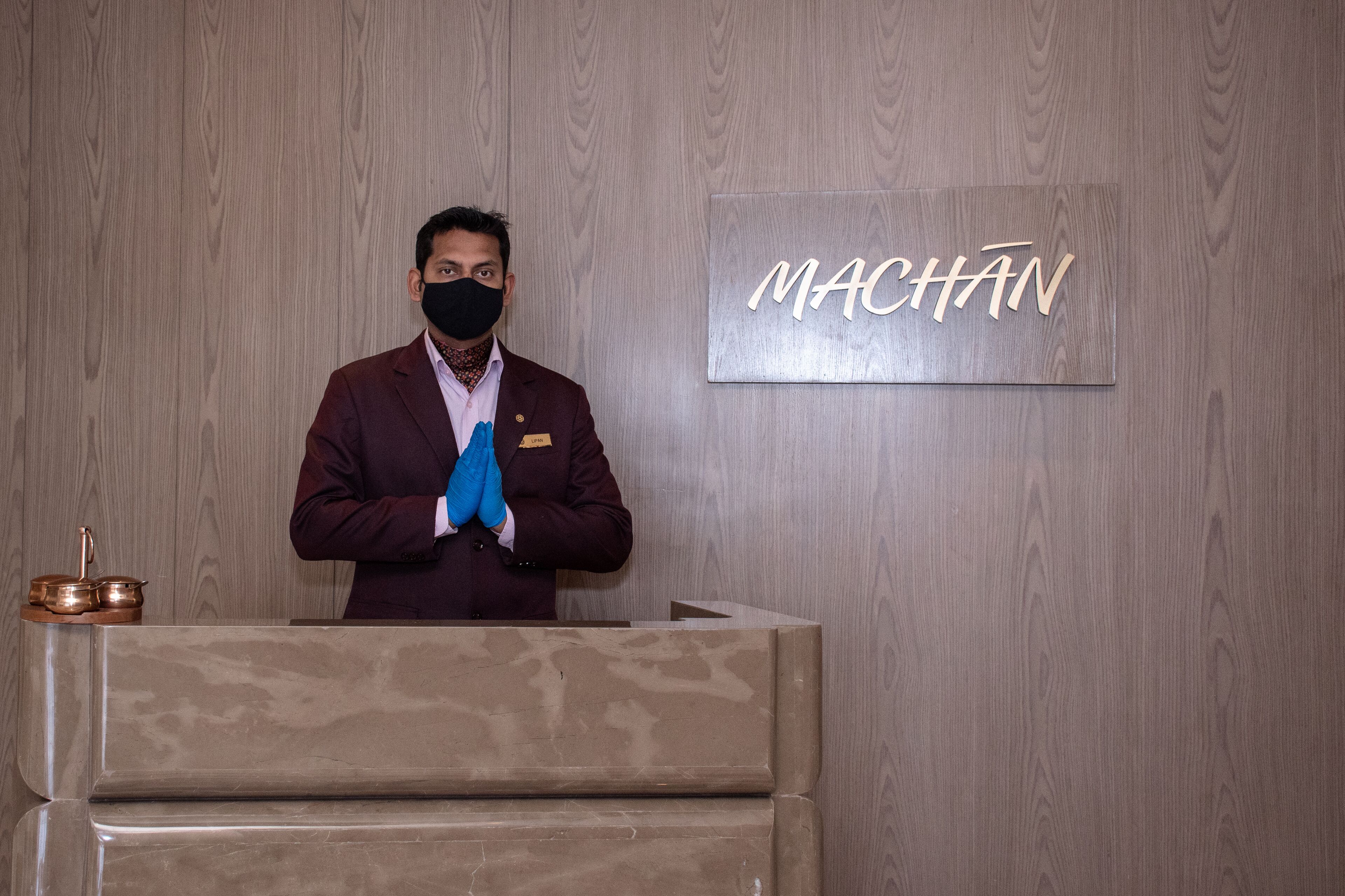 Une personne se tient derrière un comptoir d’accueil, et un panneau portant l’inscription « MACHAN » est fixé au mur derrière elle.