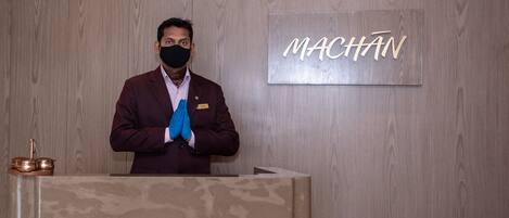 Une personne se tient derrière un comptoir d’accueil, et un panneau portant l’inscription « MACHAN » est fixé au mur derrière elle.