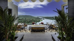 A terrace with a dining area, potted plants, and a view of a river and hills. - Taj Lakefront Bhopal (Bhopal)