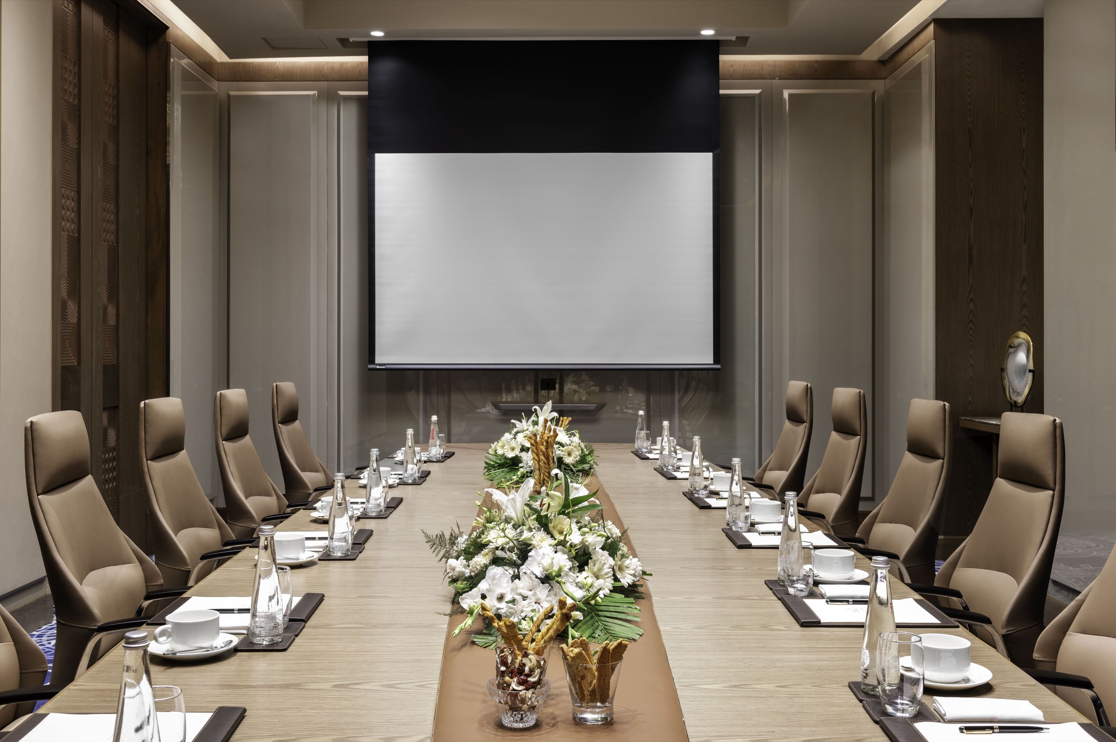 Une salle de conférence avec une longue table, des chaises de couleur beige et un grand écran à l’avant.