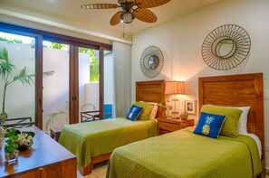 A room with two beds, a wooden table, a ceiling fan, and a view of the outdoors.