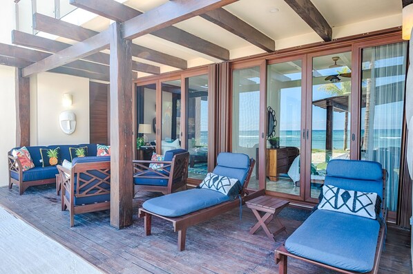 A covered outdoor seating area with wooden furniture and glass doors leading to a beach view.