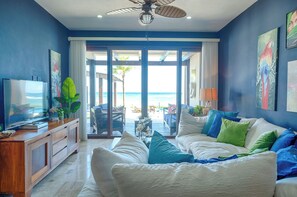 A modern living room with a large TV, a sofa with colorful cushions, and a view of the beach through large glass doors.