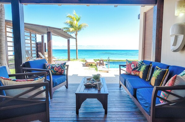 A covered outdoor seating area with a view of the ocean, featuring blue sofas and wooden chairs.