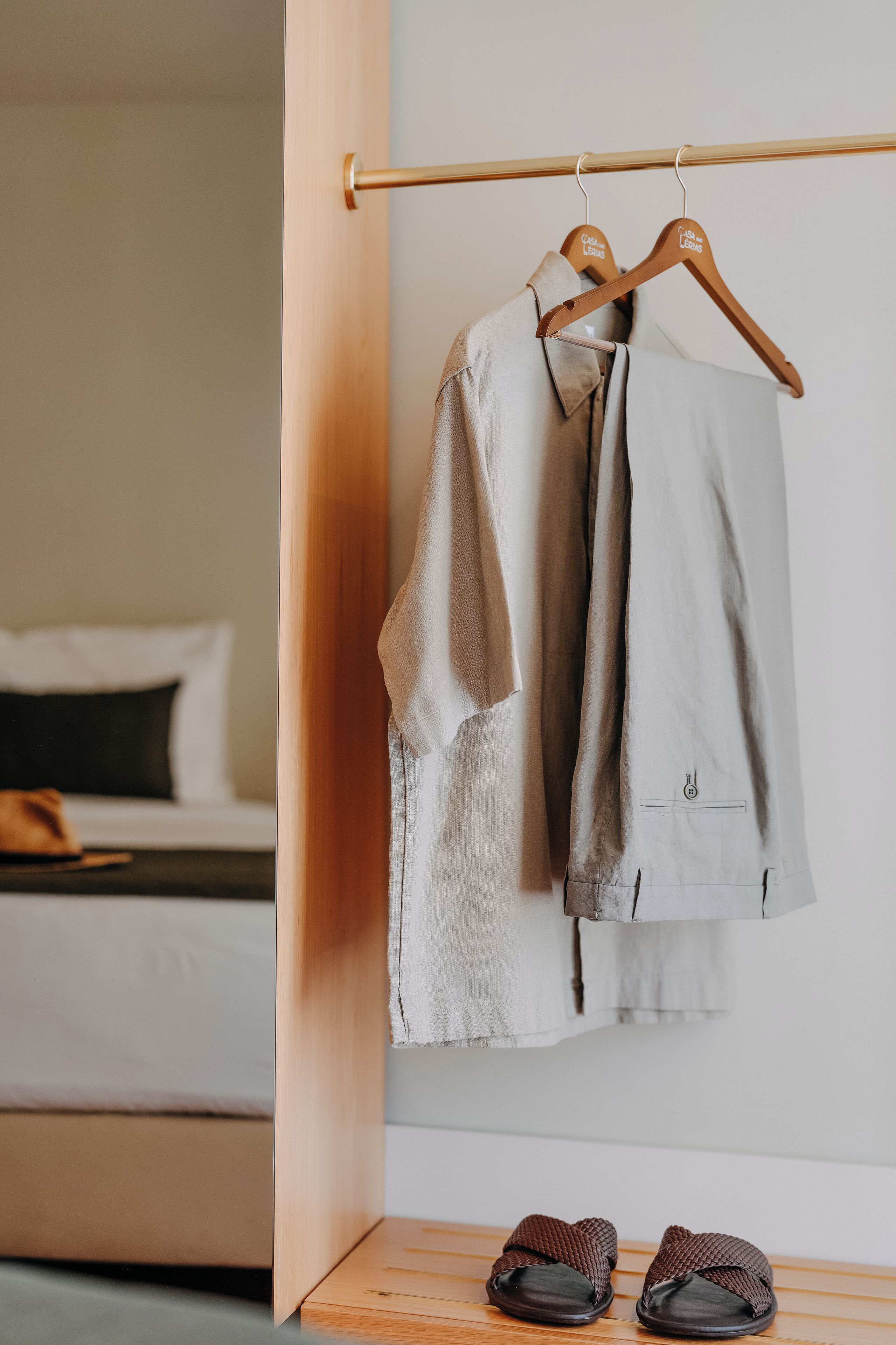 A hotel room with a bed, a wooden shelf, and a clothing rack with a shirt and slippers.
