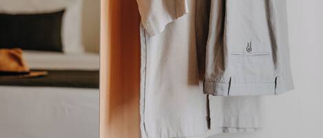 A hotel room with a bed, a wooden shelf, and a clothing rack with a shirt and slippers.