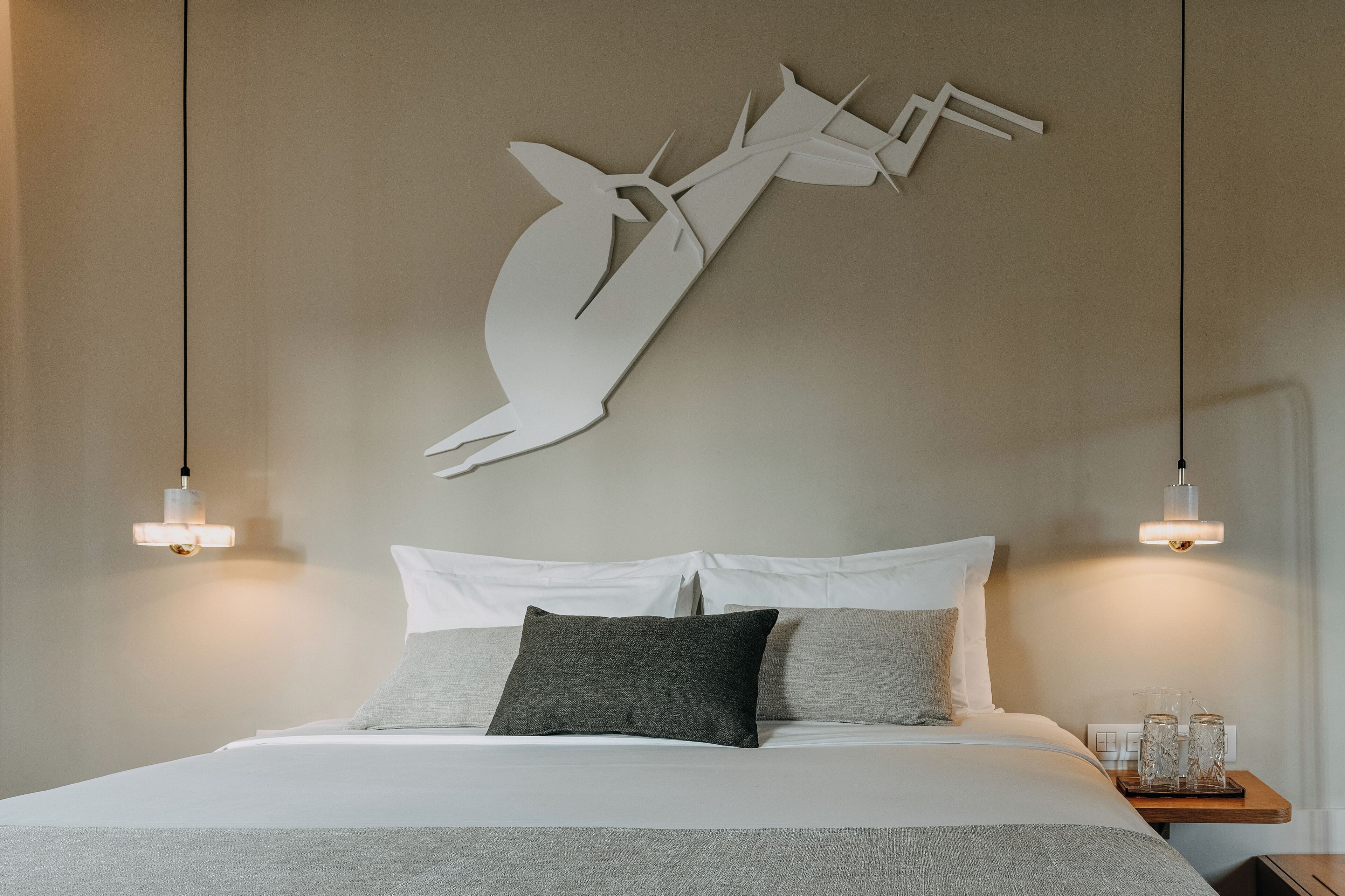 Una cama bien tendida con ropa de cama blanca y gris, una obra de arte abstracto en la pared y lámparas de noche.