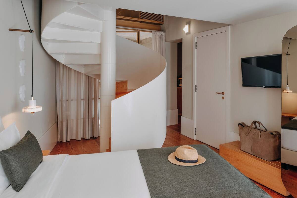 Una habitación de hotel moderna con una escalera curva, una cama con una manta verde, un sombrero sobre la cama y un televisor montado en la pared.