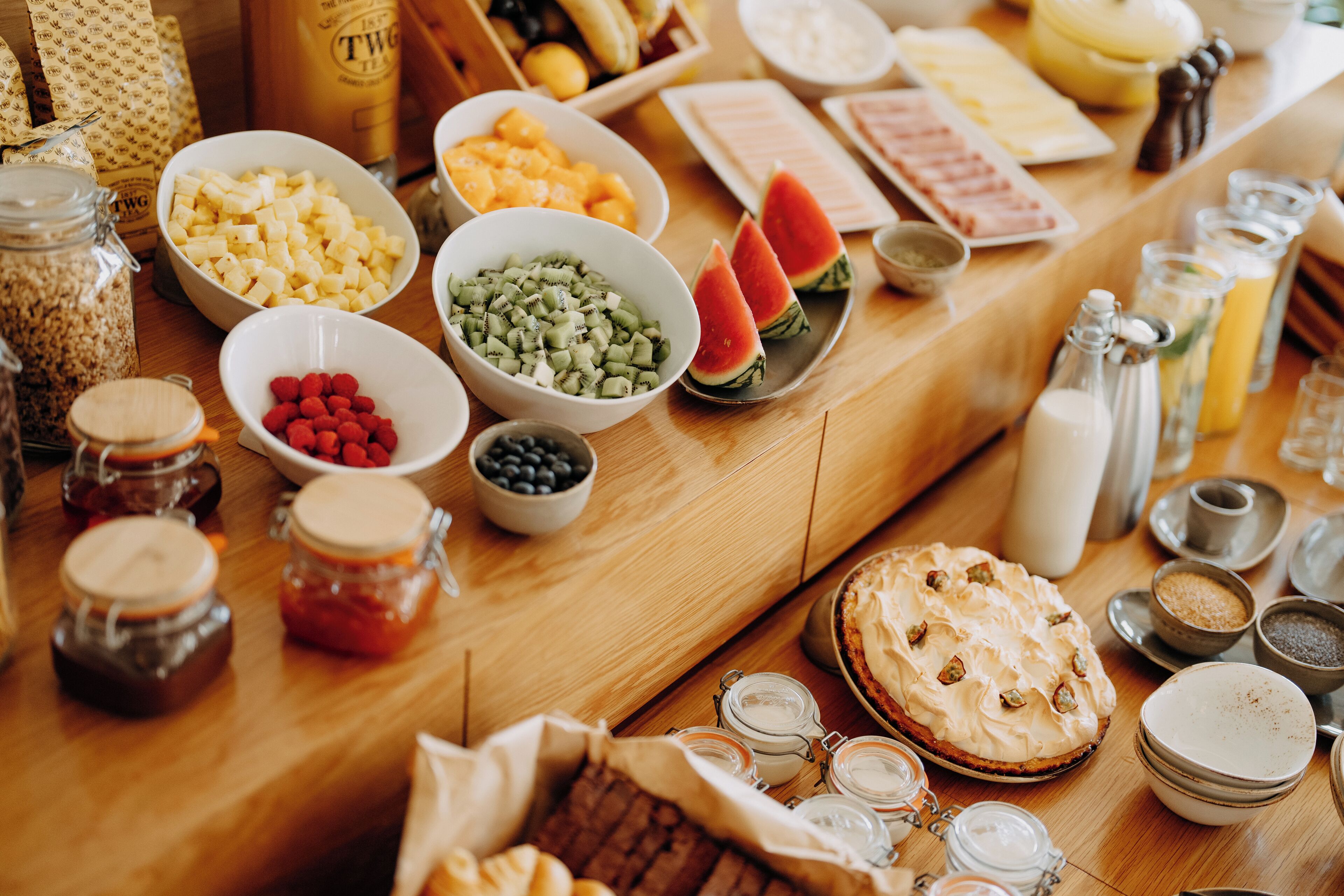 A table with various food items and condiments, including bowls of fruit, jars of preserves, and a pie.