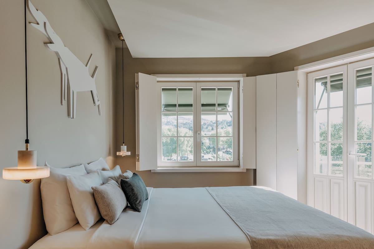 Un dormitorio con cama, almohadas, lámpara colgante, ventana con vista y una puerta.