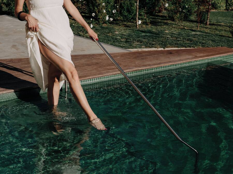 Una persona con un vestido blanco de pie junto a la piscina, sosteniendo un utensilio para limpiar la piscina.