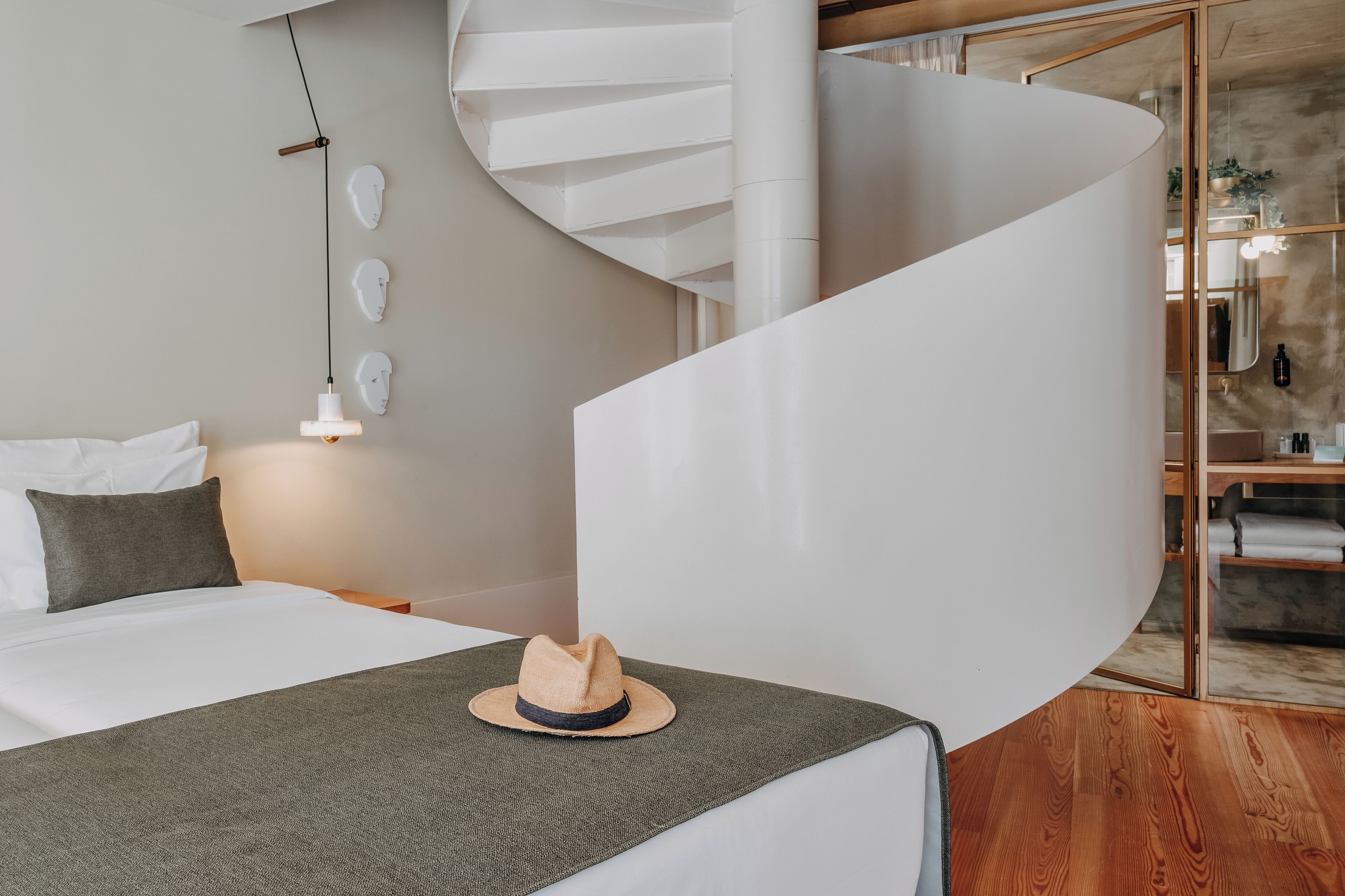 Una habitación de hotel moderna con una escalera espiral, una cama con una manta gris y un sombrero sobre la cama.