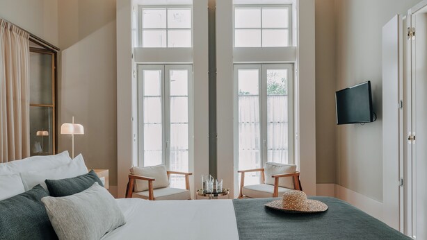 Una habitación de hotel con una cama grande, dos sillas, un televisor y vistas a través de puertas dobles.