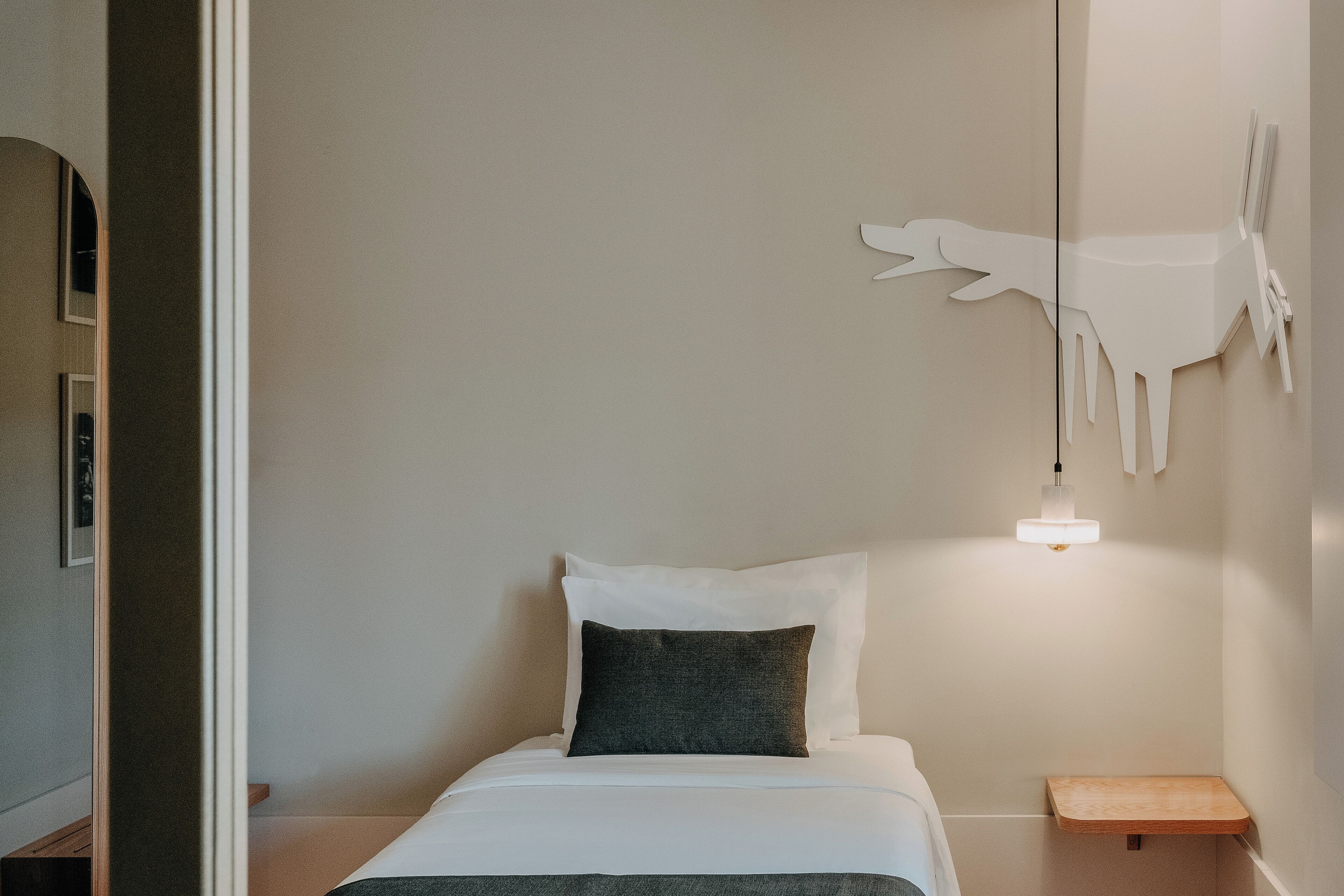 Una cama bien tendida con ropa de cama blanca y un cojín oscuro, una mesita de noche con una lámpara y una obra de arte colgada en la pared.