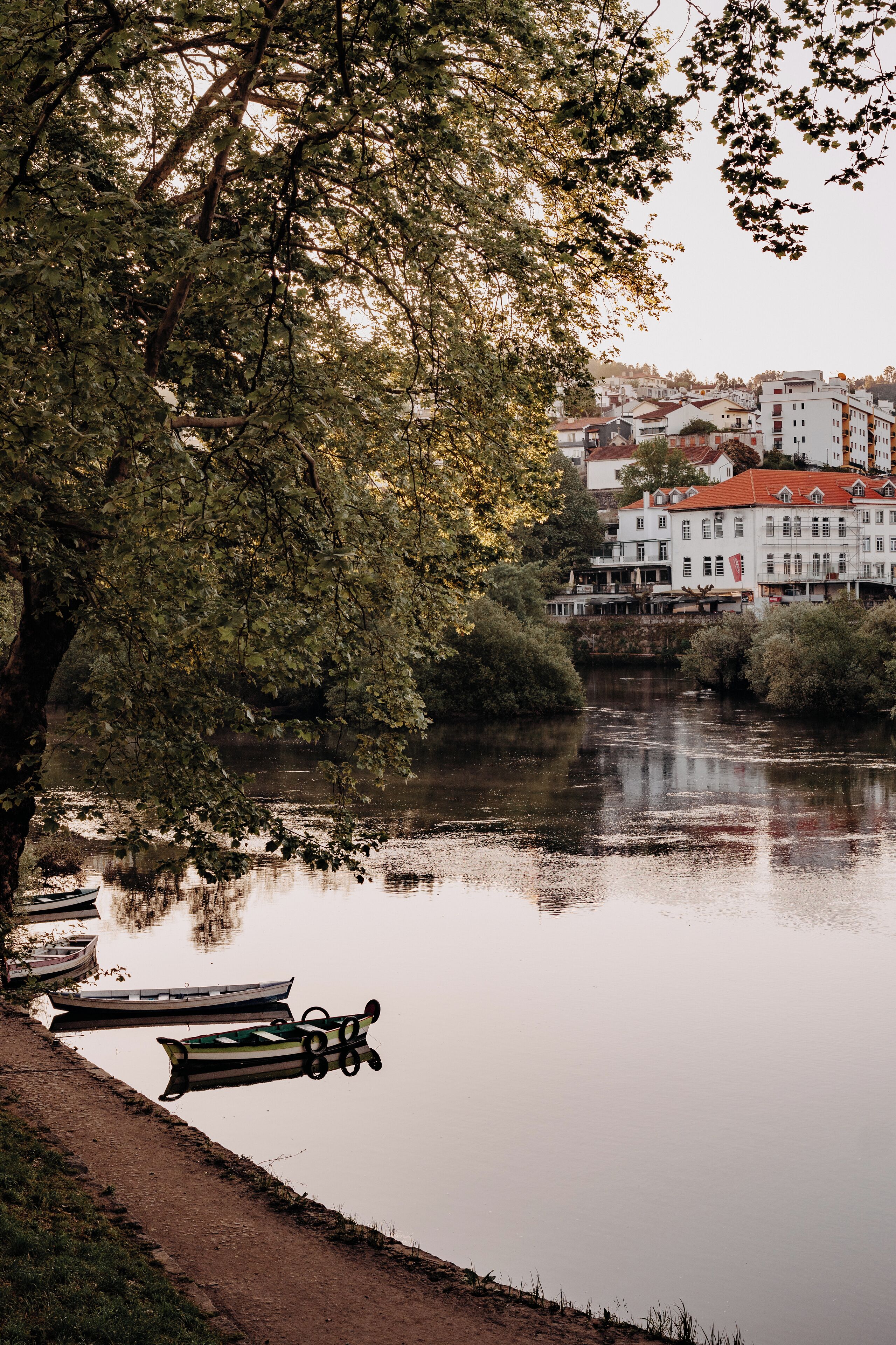 Un río tranquilo con barcos amarrados a la orilla, rodeado de árboles y edificios al fondo.