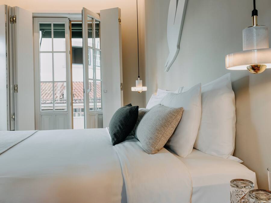 A neatly made bed with white bedding and a variety of pillows, a bedside table with a glass container, and a view of a building through an open door.