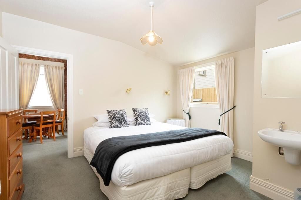 A bedroom with a large bed, a bedside table, a hanging light fixture, a window with curtains, and a bathroom with a sink.