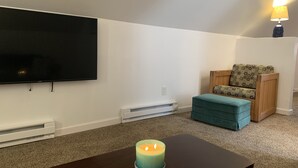 Una sala de estar con un televisor de pantalla plana, un reposapiés color turquesa, una silla de madera y una vela encendida sobre una mesa.