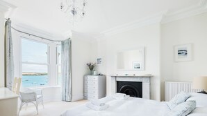 A bedroom with a large bed, a dresser, a chair, a fireplace, and a view of the sea. - The Terrace 2 (Wadebridge)