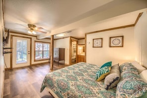 A bedroom with a patterned bedspread, a ceiling fan, and a view of the backyard through a door. - RR 1300 Hondo's River Haus (New Braunfels)