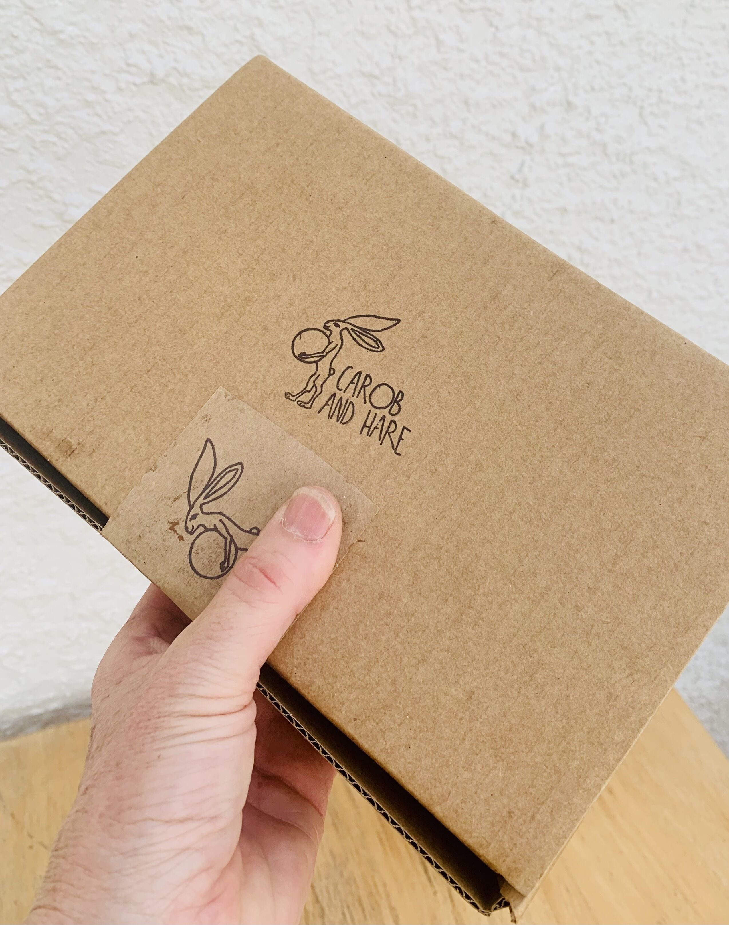 Une main tenant une boîte en carton marron ornée d’une illustration de lapin et portant l’inscription « CAROB AND HARE ».