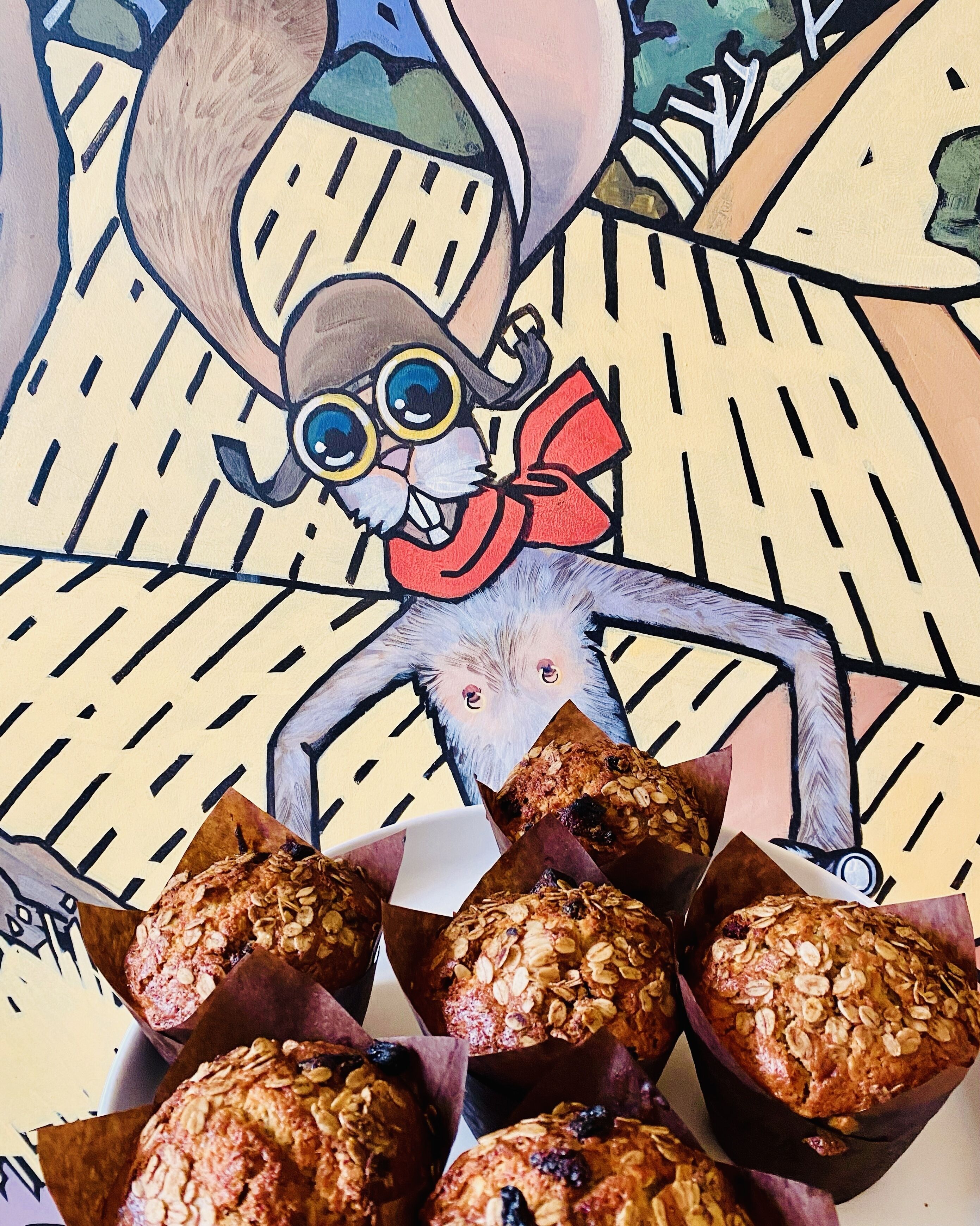 A character with a large hat and glasses sitting behind muffins in a box.
