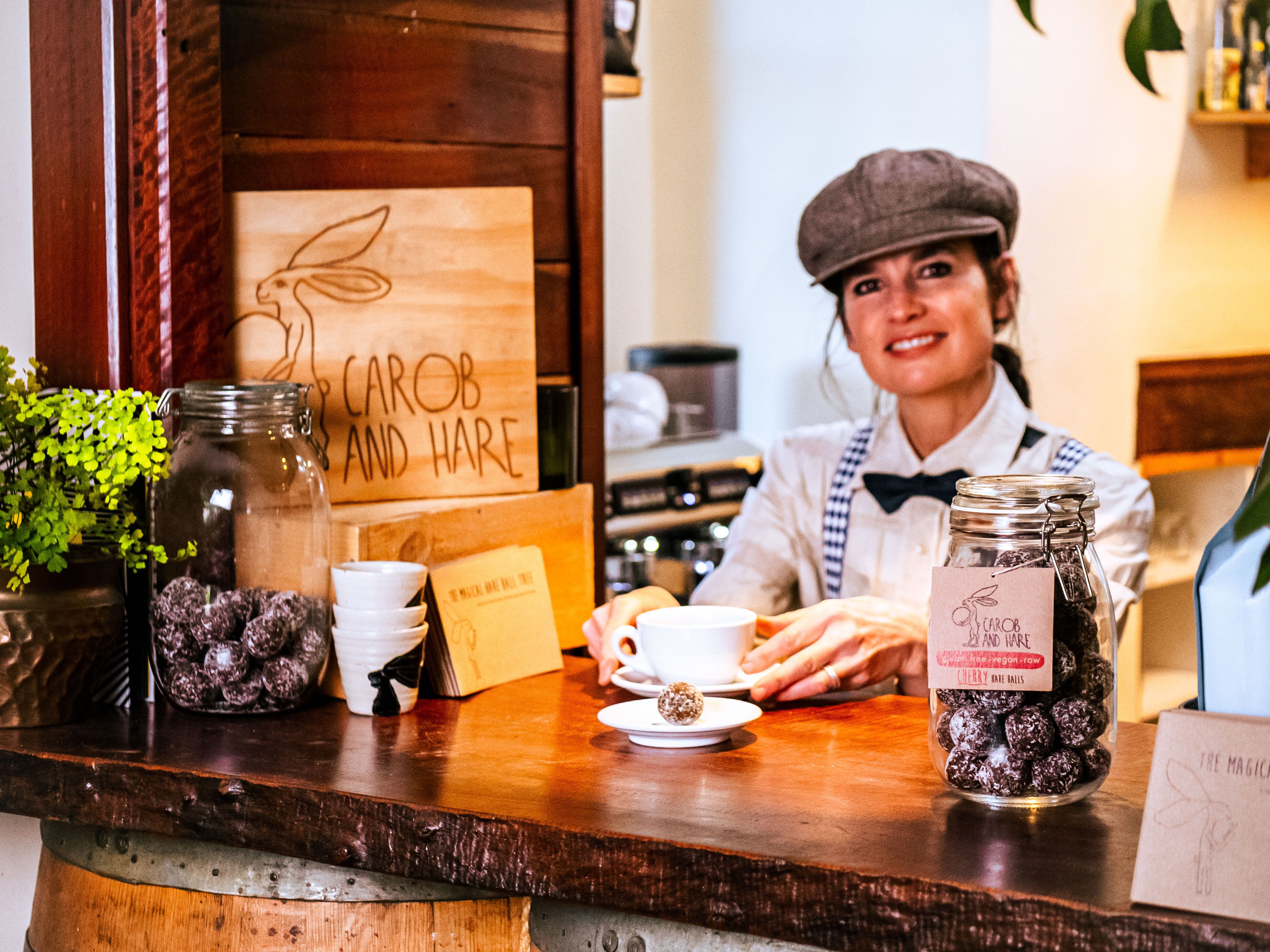 A barista at a counter with a wooden sign reading "CAROB AND HARE," jars of chocolate balls, a coffee cup, and a menu.