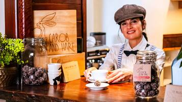 A barista at a counter with a wooden sign reading "CAROB AND HARE," jars of chocolate balls, a coffee cup, and a menu.