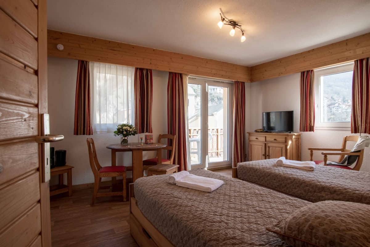 A room with two beds, a TV, a table with chairs, and a balcony with curtains.