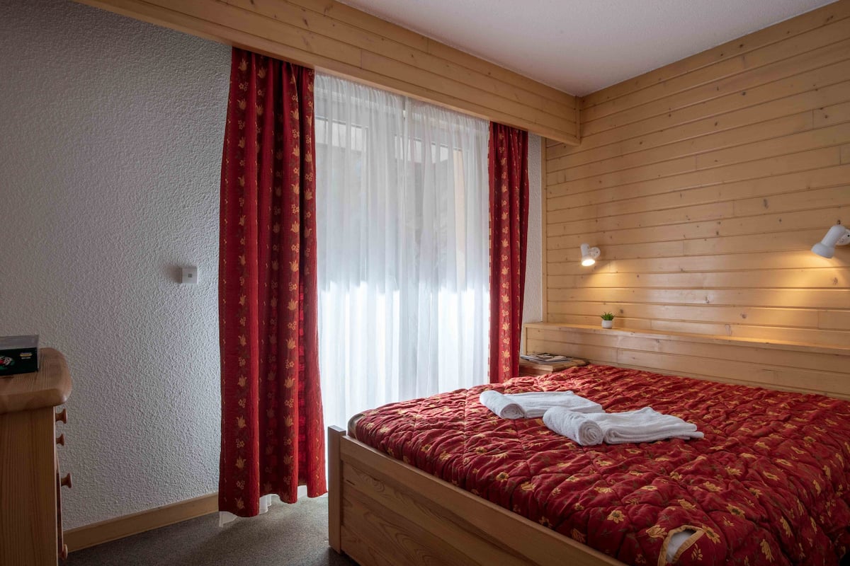 A wooden-paneled room with a bed, red curtains, and a small bedside table.