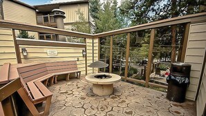 Une terrasse extérieure avec un sol en pierre, un foyer, des bancs en bois et une poubelle.