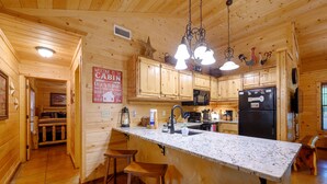 A wooden cabin interior with a kitchen featuring a marble countertop, wooden stools, and a black refrigerator.