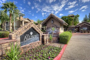 A sign for The Allison Condominiums with a brick archway, a small building with a tiled roof, and a landscaped area with flowers and palm trees.