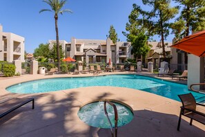 A swimming pool with lounge chairs and umbrellas, surrounded by a building with balconies and palm trees.