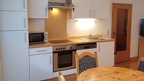 Cocina compacta con armarios blancos, microondas, horno y lavamanos. Hay una mesa de comedor de madera con sillas.