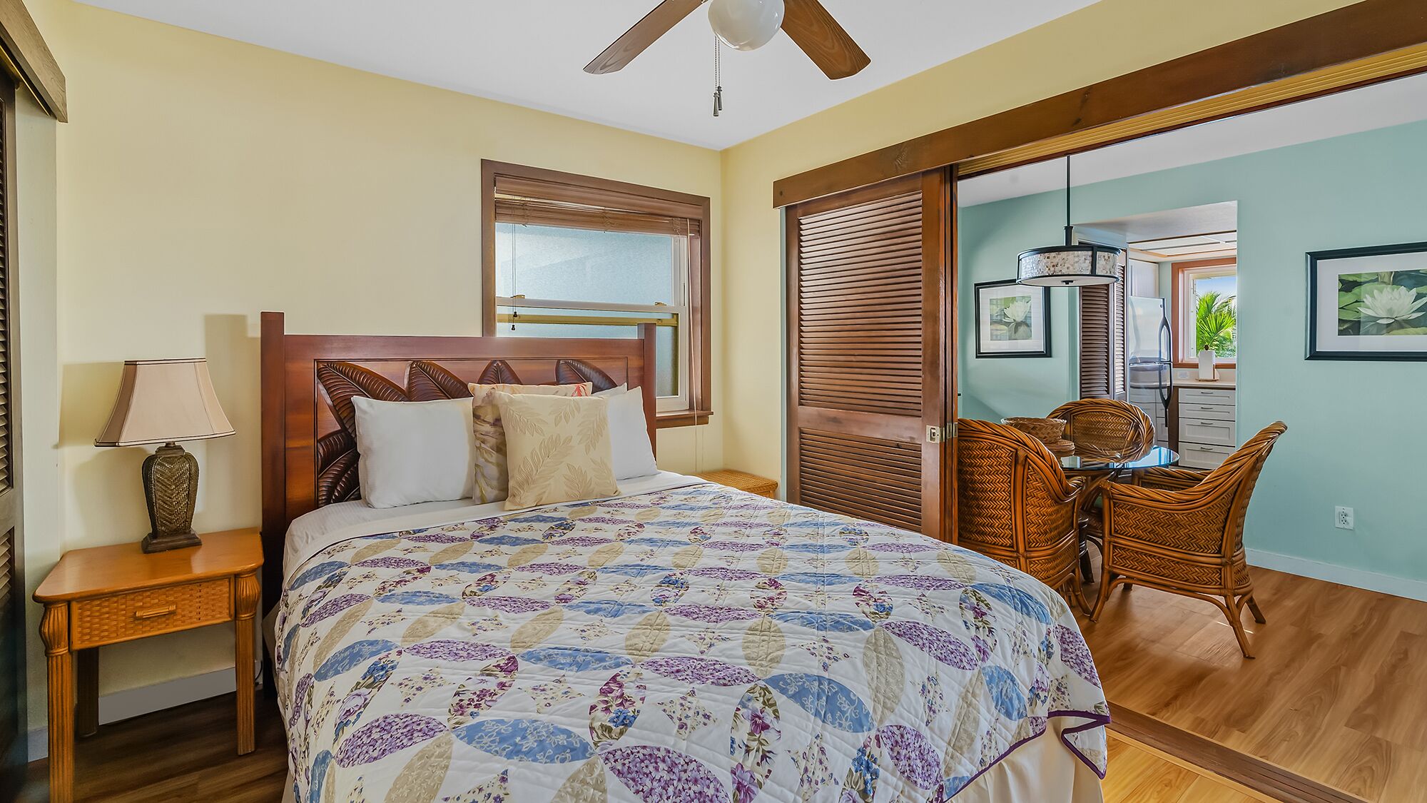 A bedroom with a bed, a nightstand, a lamp, a ceiling fan, a dining area with wicker chairs, and a view through a sliding door.