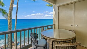A balcony with a glass table and wicker chairs, overlooking a clear blue ocean and palm trees. - Poipu Palms 303 By Parrish Kauai - oceanfront views & walk to beach (Koloa)
