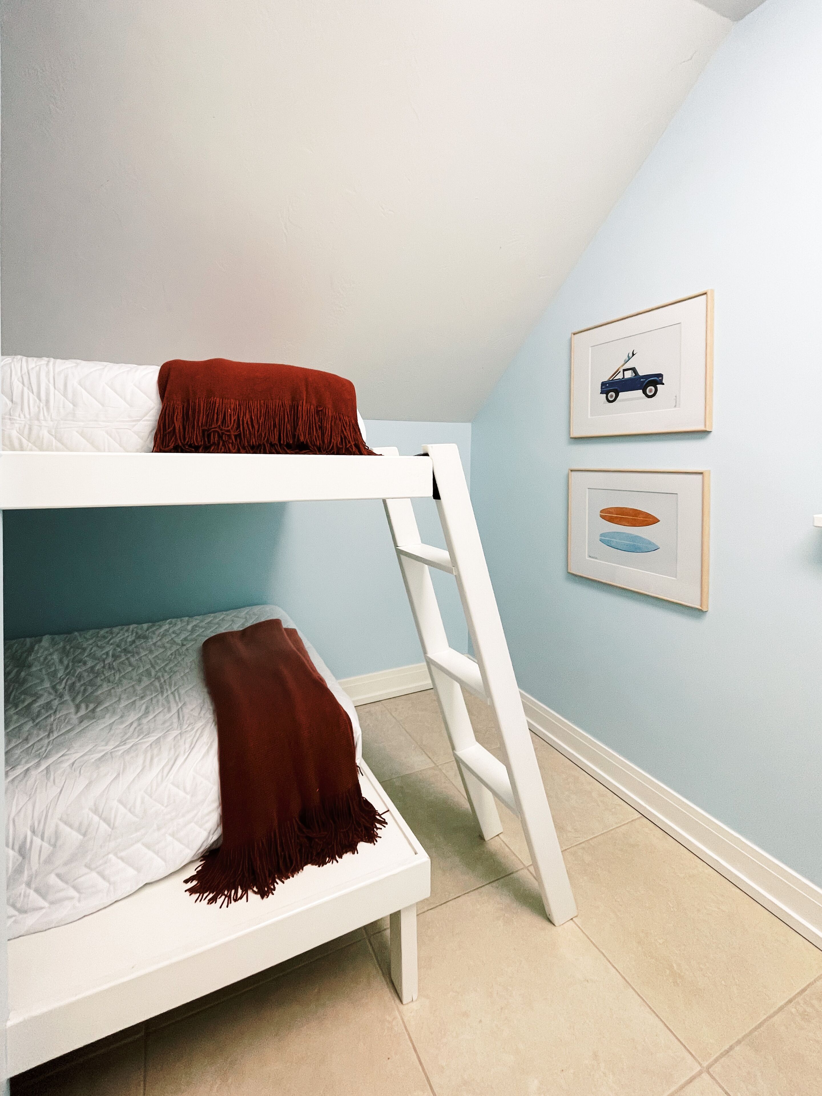 A bunk bed with a ladder, white bedding, and a brown throw blanket. There are two framed pictures on the wall.