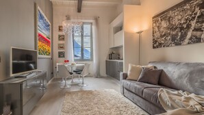 A modern living room with a grey sofa, a TV, a dining area with a table and chairs, a chandelier, and artwork on the walls. - Il Diamante del Lago (Varenna)