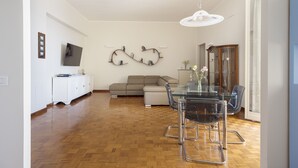Ein geräumiger Wohnbereich mit Holzboden, einem Glastisch mit Stühlen, einer Couch und einem an der Wand befestigten Fernseher.