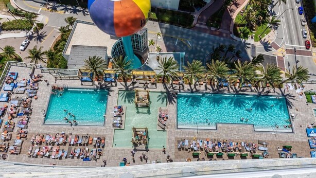 Un área de piscina en la azotea con dos piscinas, sillas de descanso y palmeras.