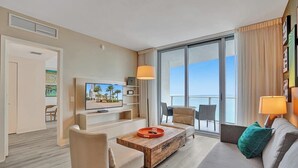 Habitación de hotel moderna con un balcón grande, un televisor de pantalla plana, una mesa de centro de madera y un sofá con un cojín azul turquesa.